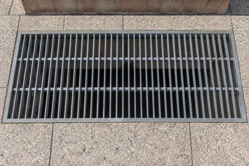 Grille de ventilation encastrée dans une dalle piétonne, vide du parking visible en dessous