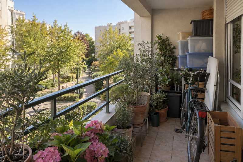 Loggia encombrée d'objets accumulés, garde-corps métallique peint, mail planté en contrebas